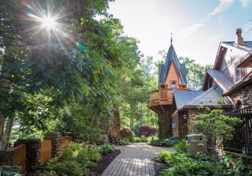 A sunlit pathway leads to a charming stone house with a turret, surrounded by lush greenery and vibrant flowers.