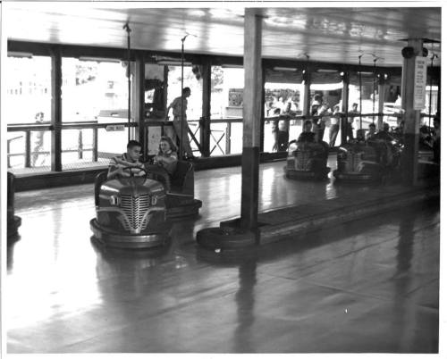 A black and white photo of a bumper car ride with two children inside a car, surrounded by empty cars and a bright interior.