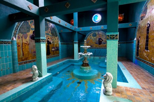 A serene indoor pool area with turquoise walls, a central fountain, and decorative murals, featuring lion statues.
