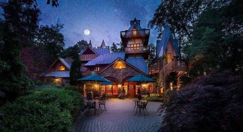 A charming stone house illuminated under a starry night sky, with a full moon and outdoor seating areas.
