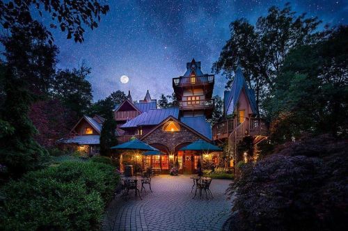 A charming stone house with a tower, illuminated under a starry night sky and a bright moon, surrounded by trees.