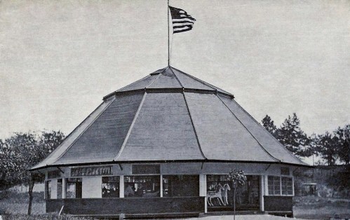 A vintage octagonal building with a flag on top, surrounded by trees, featuring a horse statue outside.