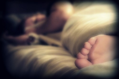 A close-up of a child's foot resting on a bed, with a blurred figure of an adult in the background.