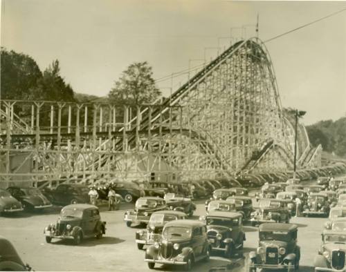 A vintage photo of a wooden roller coaster with classic cars parked in the foreground.