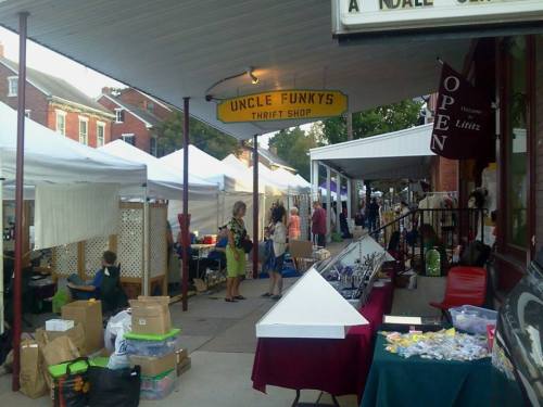 A bustling outdoor market scene with tents, vendors, and shoppers near Uncle Funky's Thrift Shop.