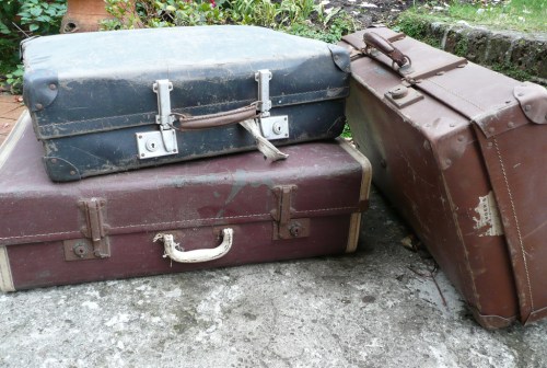 Three vintage suitcases in varying colors and conditions, resting on a stone surface with greenery in the background.