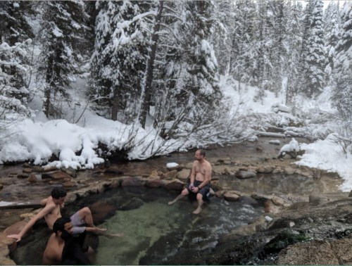 A group of people relaxes in a natural hot spring surrounded by snow-covered trees and a winter landscape.