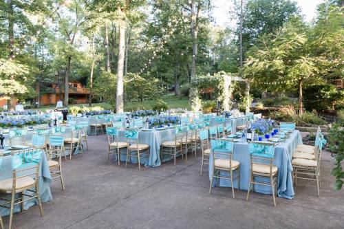 Outdoor event setup with tables dressed in blue and gold, surrounded by greenery and floral arrangements.