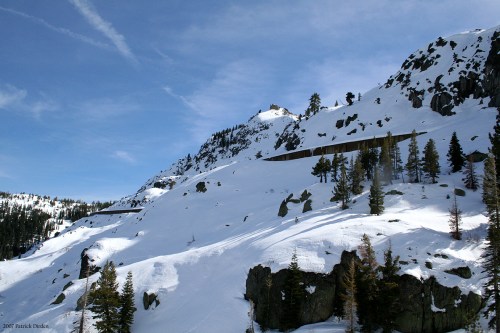 Snow-covered mountain landscape with rocky outcrops and scattered pine trees under a clear blue sky.