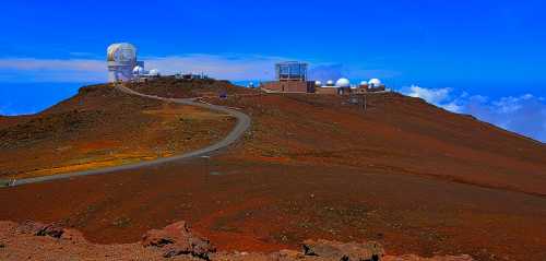 A panoramic view of a mountain summit with observatories and a winding road under a clear blue sky.