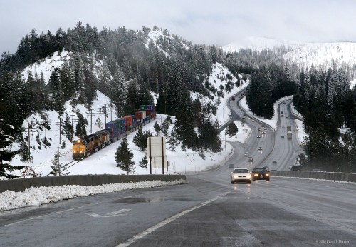 A train travels alongside a snowy highway, with cars navigating the winding road through a winter landscape.