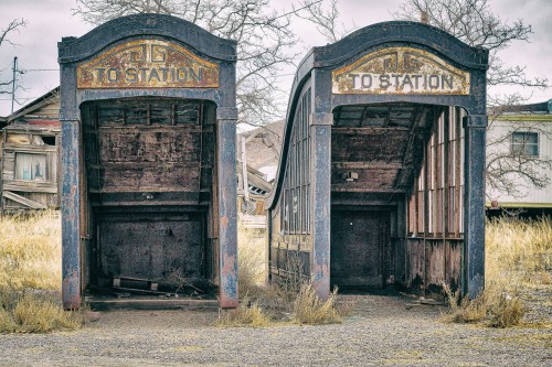 Two vintage, abandoned bus shelters with "TO STATION" signs, surrounded by overgrown grass and dilapidated buildings.