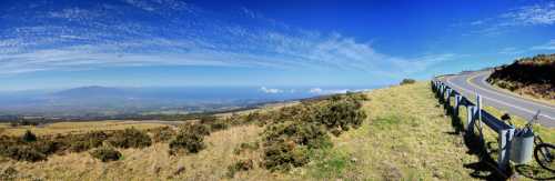 Panoramic view of a scenic landscape with rolling hills, a winding road, and a clear blue sky.