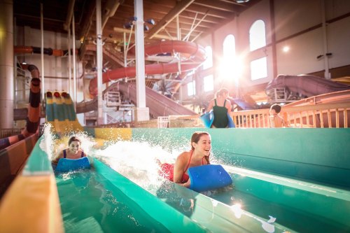 Two people enjoy a water slide at an indoor water park, splashing through the water with bright sunlight streaming in.