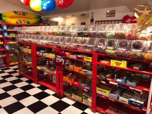 A colorful candy store display with jars of sweets and various candy boxes on red shelves against a black and white checkered floor.