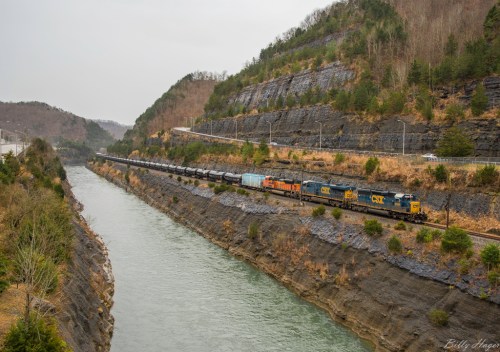 A freight train travels along a river, flanked by steep, rocky hills in a scenic landscape.
