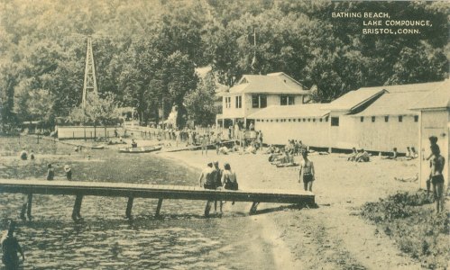 Historic photo of Bathing Beach at Lake Compounce, Bristol, Connecticut, featuring people by the water and nearby buildings.