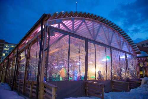 A large, illuminated tent with transparent walls, set against a twilight sky, surrounded by snow.