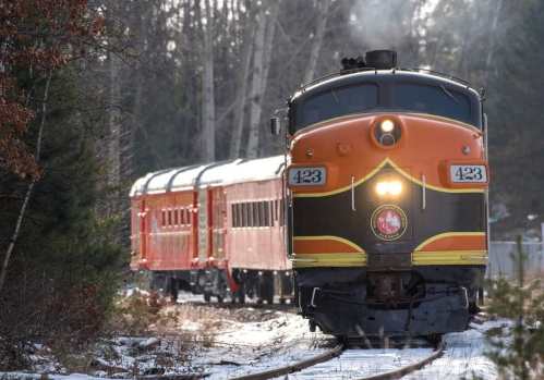 A vintage orange and black train with passenger cars travels along a snowy track in a wooded area.