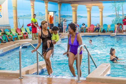 Two women enter a pool area, one carrying a child, while others enjoy the water and relax on colorful chairs.