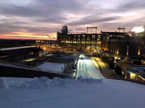 A snowy landscape at dusk, with a stadium in the background and a colorful sunset illuminating the sky.