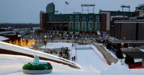 A snowy hill with a green sled in the foreground, and a stadium in the background under a gray sky.