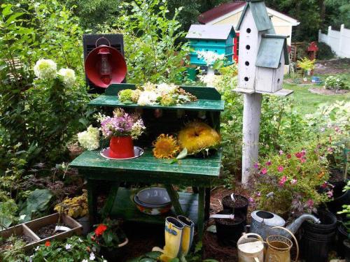A vibrant garden scene featuring a green shelf with flowers, a sunflower, and colorful birdhouses surrounded by lush plants.