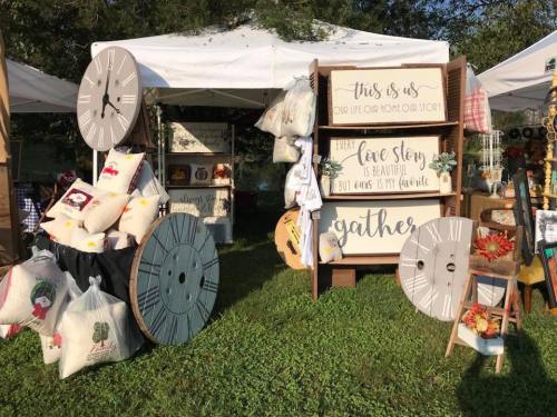 A craft booth featuring decorative signs, pillows, and large clock displays under a white tent in a grassy area.