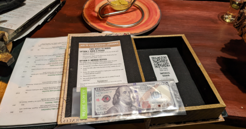 A wooden box containing a menu, a QR code, and a $100 bill on a table.