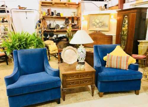 Two blue upholstered chairs flanking a wooden side table with a lamp, surrounded by vintage decor in a cozy setting.