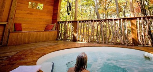 A person relaxes in a hot tub on a wooden deck surrounded by trees in a serene, natural setting.