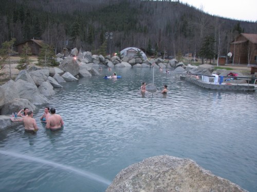 A natural hot spring with people relaxing in the water, surrounded by rocks and trees in a serene outdoor setting.