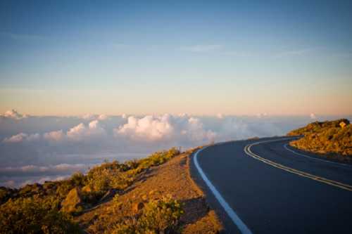 A winding road curves through a mountainous landscape, with clouds below and a colorful sunset in the sky.