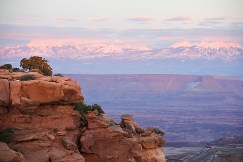 A scenic view of rocky cliffs with snow-capped mountains in the background, bathed in soft pink light at sunset.