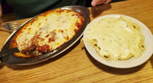 A sizzling dish of cheesy baked meat with marinara sauce next to a plate of creamy fettuccine alfredo.