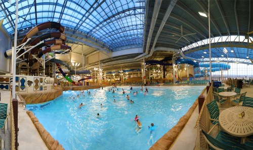 Indoor water park featuring a large wave pool, water slides, and numerous people enjoying the water. Bright, spacious environment.