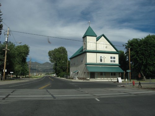 A large, green-roofed church stands at a street corner, surrounded by trees and mountains under a cloudy sky.