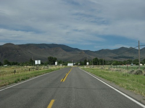 A straight road stretches into the distance, flanked by green fields and mountains under a cloudy sky.