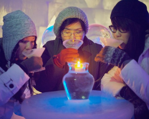 Three people in winter attire sipping drinks from glasses, with a glowing candle on an ice table in a colorful ice setting.