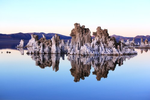 Tufa formations rise from a calm lake, reflecting in the water under a pastel sky with distant mountains.