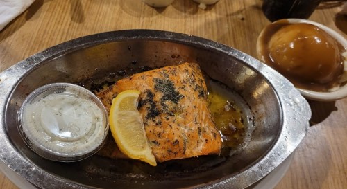 A plated salmon fillet with herbs, a lemon slice, and a side of sauce, next to a scoop of dessert on a wooden table.
