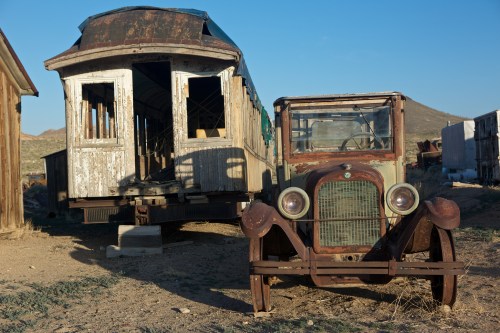 An old, rusty car parked in front of a dilapidated train car in a desert setting.