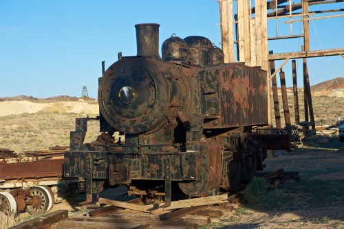 An old, rusted steam locomotive sits on wooden tracks in a desert landscape, with a clear blue sky in the background.