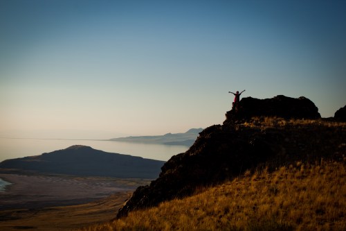 A person stands on a rocky outcrop, arms raised, overlooking a serene landscape with mountains and water in the distance.