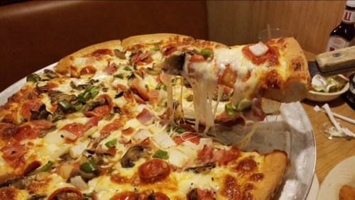 A close-up of a large pizza with melted cheese, pepperoni, vegetables, and a slice being lifted with gooey cheese stretching.