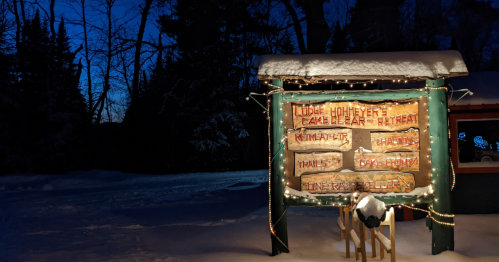 A snowy sign for "Lodge Homeyer" illuminated with lights, surrounded by trees at dusk.