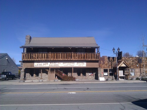 A rustic building with a sign reading "Golden Saloon," featuring a balcony and stone accents, set along a street.