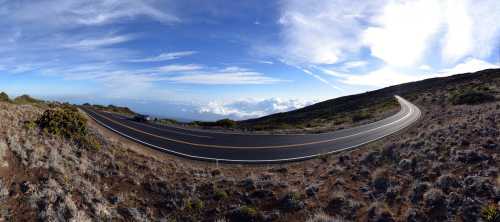 A winding road curves through a mountainous landscape under a blue sky with scattered clouds.