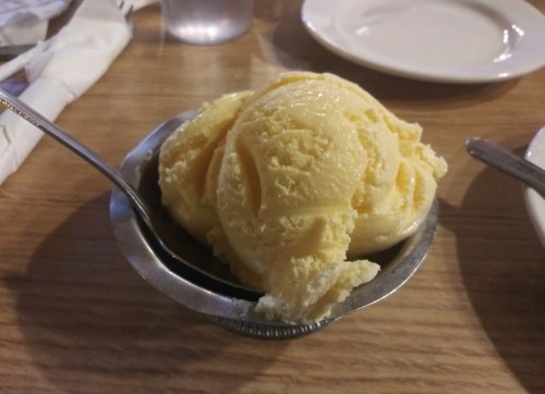A silver bowl filled with two scoops of yellow ice cream on a wooden table.