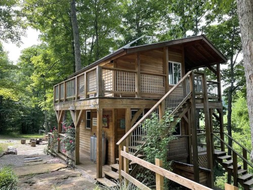 A wooden cabin elevated on stilts, surrounded by trees, with a staircase and a deck overlooking a natural setting.